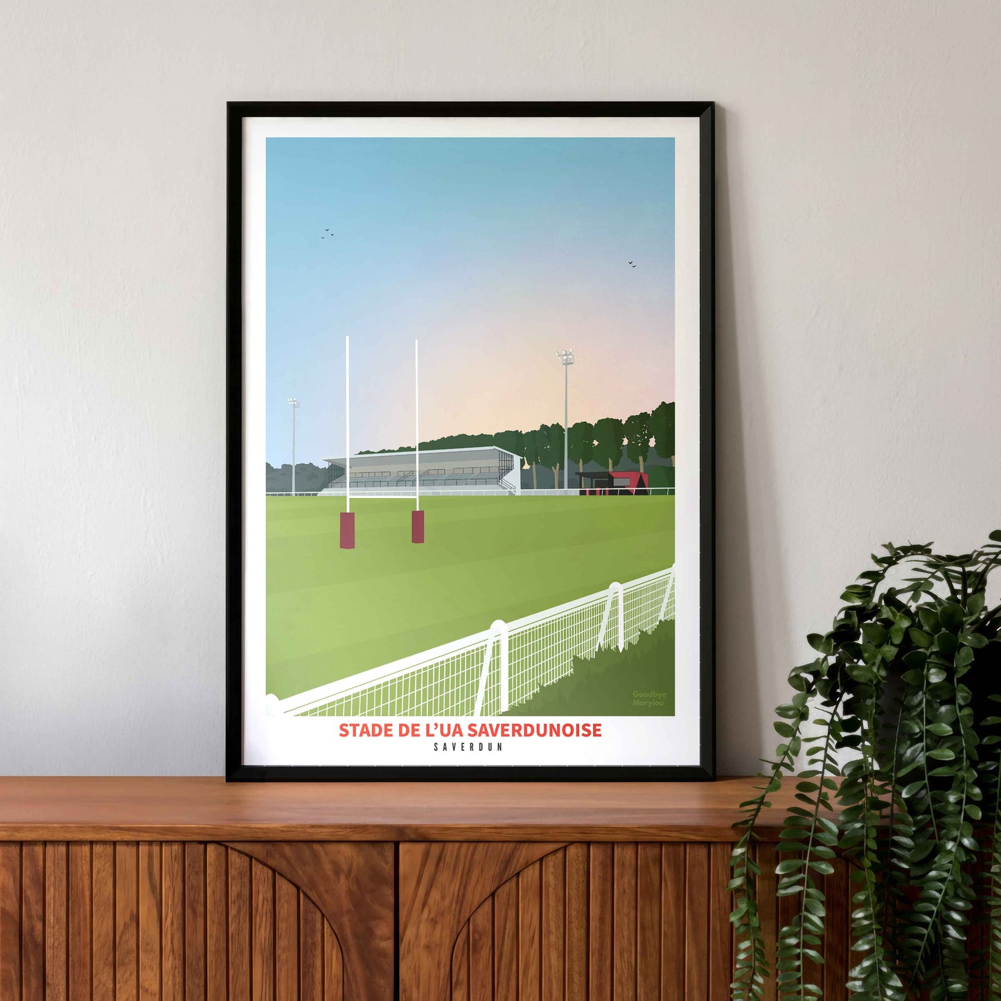 Mise en situation de l'affiche du stade de rugby de Saverdun dans un cadre noir, posée sur un buffet en bois vintage avec une plante verte tombante.