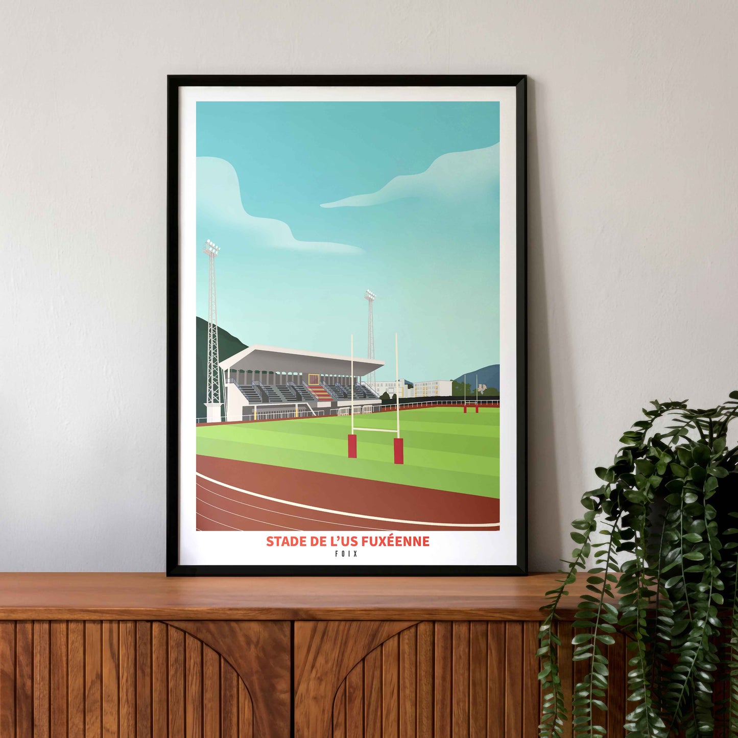 Mise en situation de l'affiche du stade Jean-Noël Fondère (Foix) dans un cadre noir, posée sur un buffet en bois vintage avec une plante tombante.