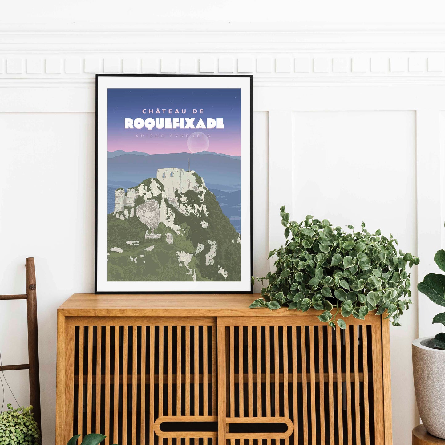 Affiche du château de Roquefixade dans un cadre noir, posée sur un buffet en bois à lattes avec une plante verte.