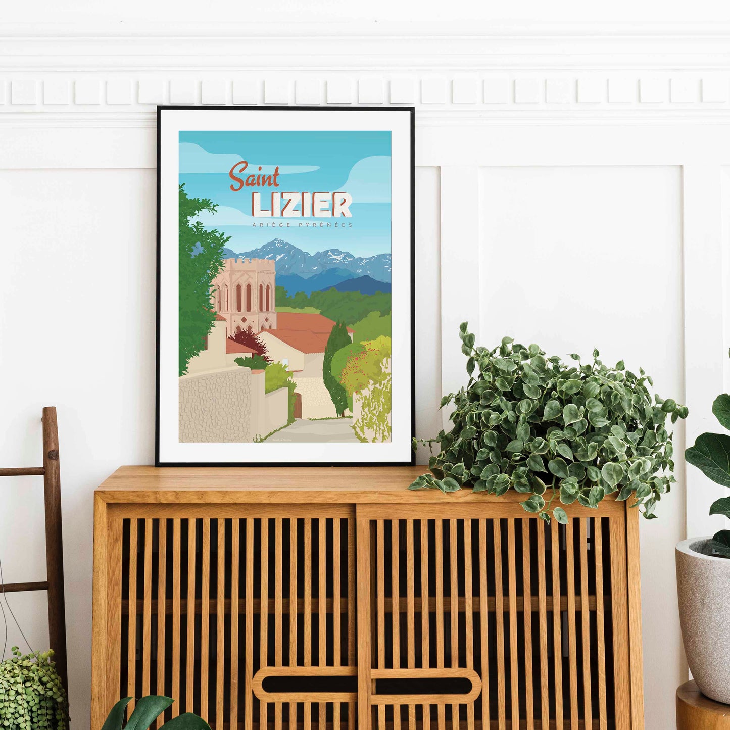 Affiche de Saint-Lizier dans un cadre noir élégant, posée sur un buffet en bois à lattes avec une grande plante verte décorative.
