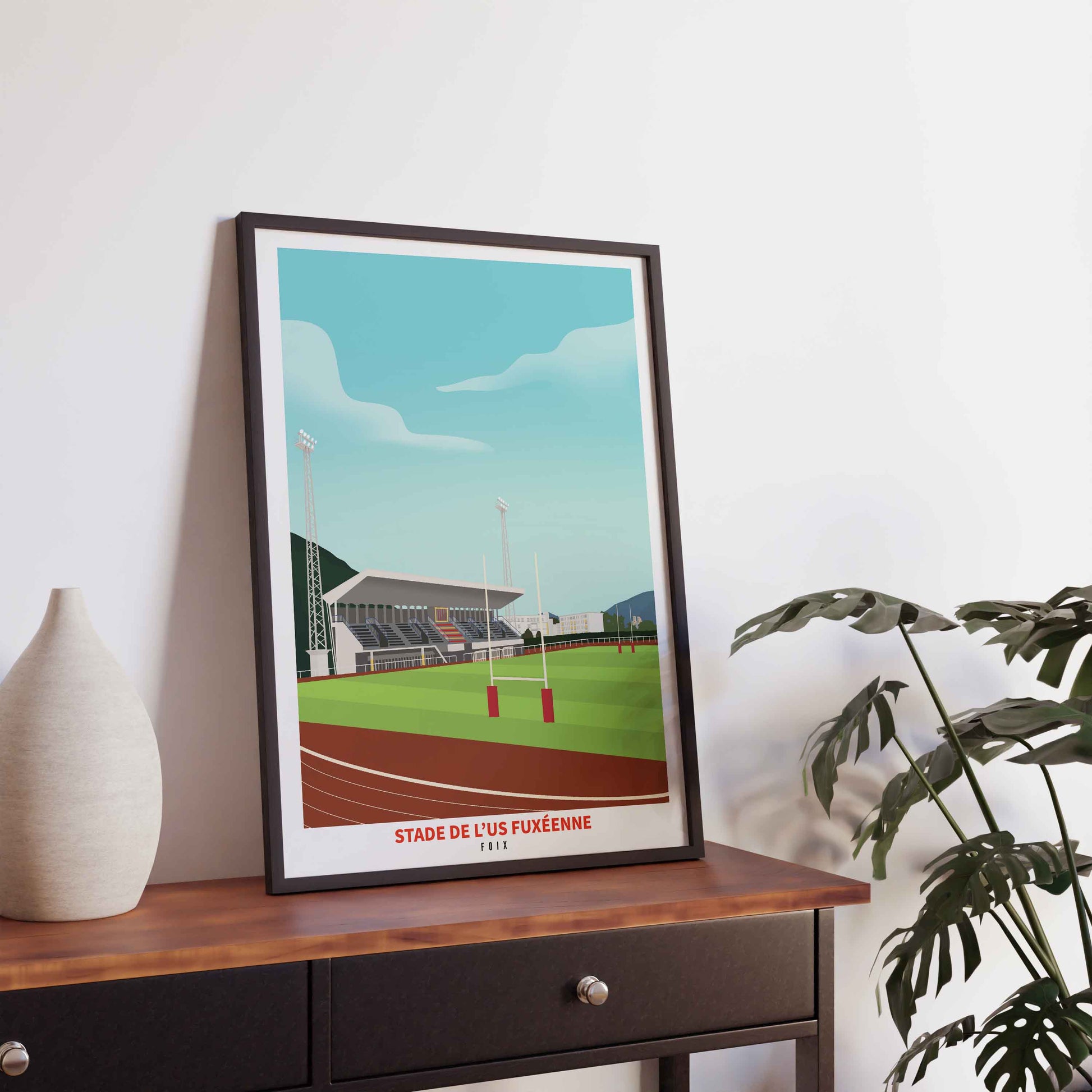 Illustration du stade de rugby de Foix dans un cadre noir fin, posée sur un meuble en bois sombre avec un vase blanc et une plante