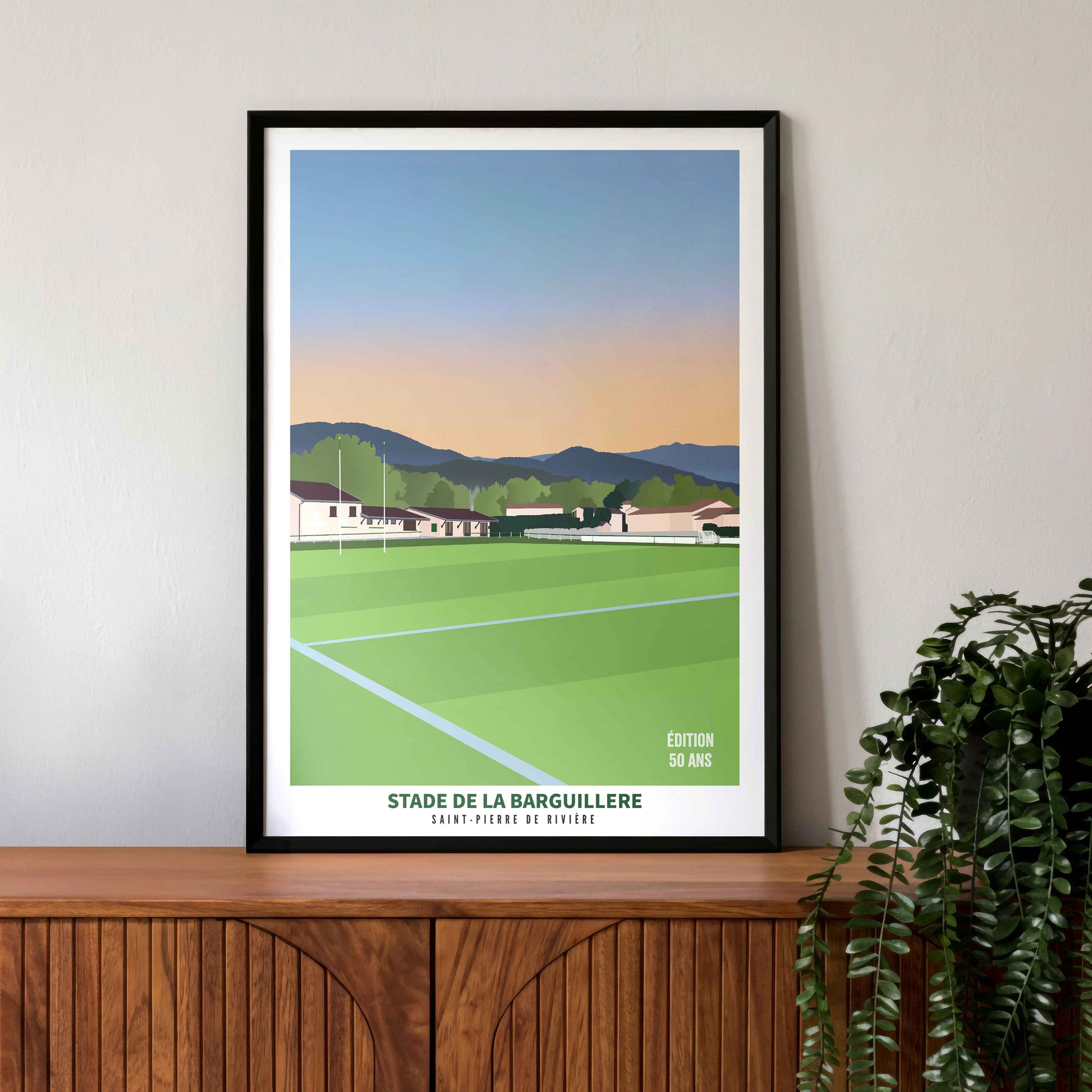 Mise en situation de l'affiche "Édition 50 ans" du stade de la Barguillère dans un cadre noir, posée sur un buffet vintage en bois.