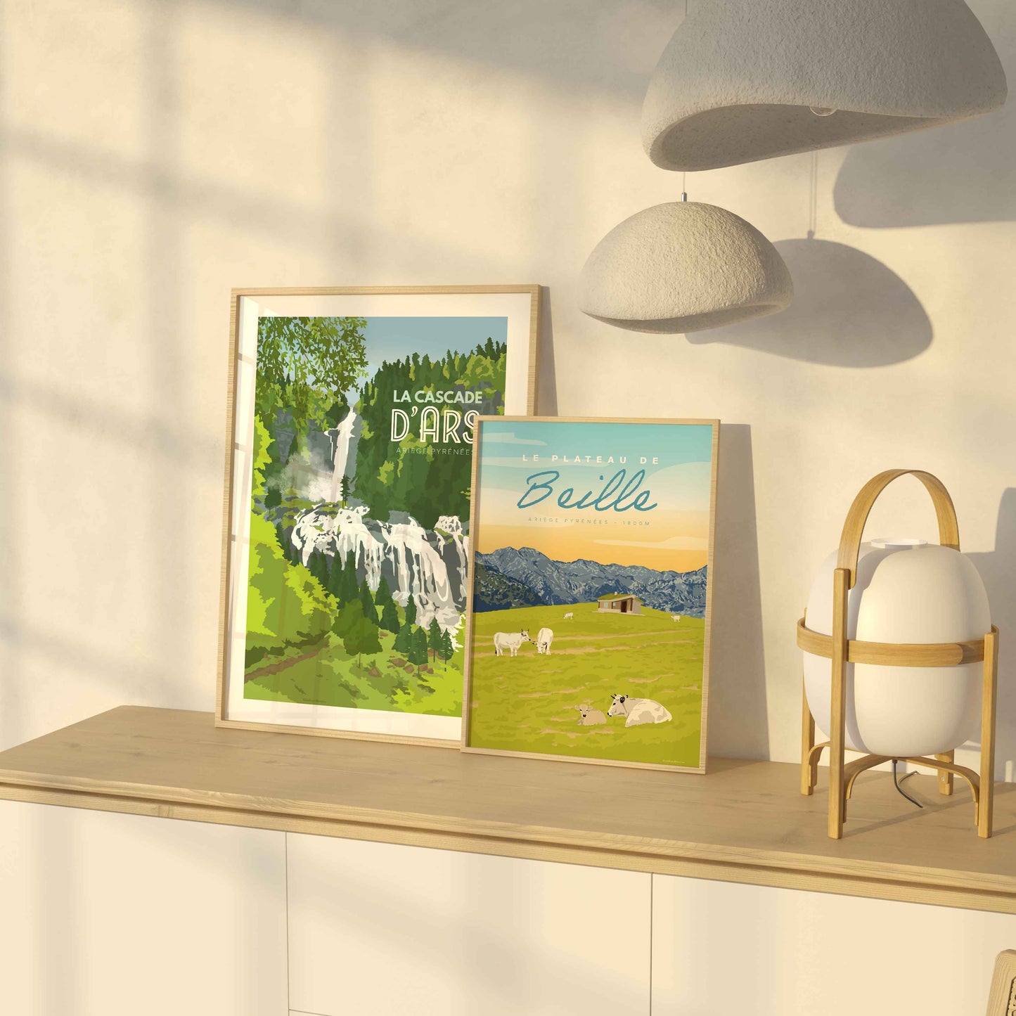 Duo d’affiches de l’Ariège (Cascade d'Ars et Plateau de Beille été) posées sur un buffet en bois clair baigné par la lumière dorée d'une fin de journée.