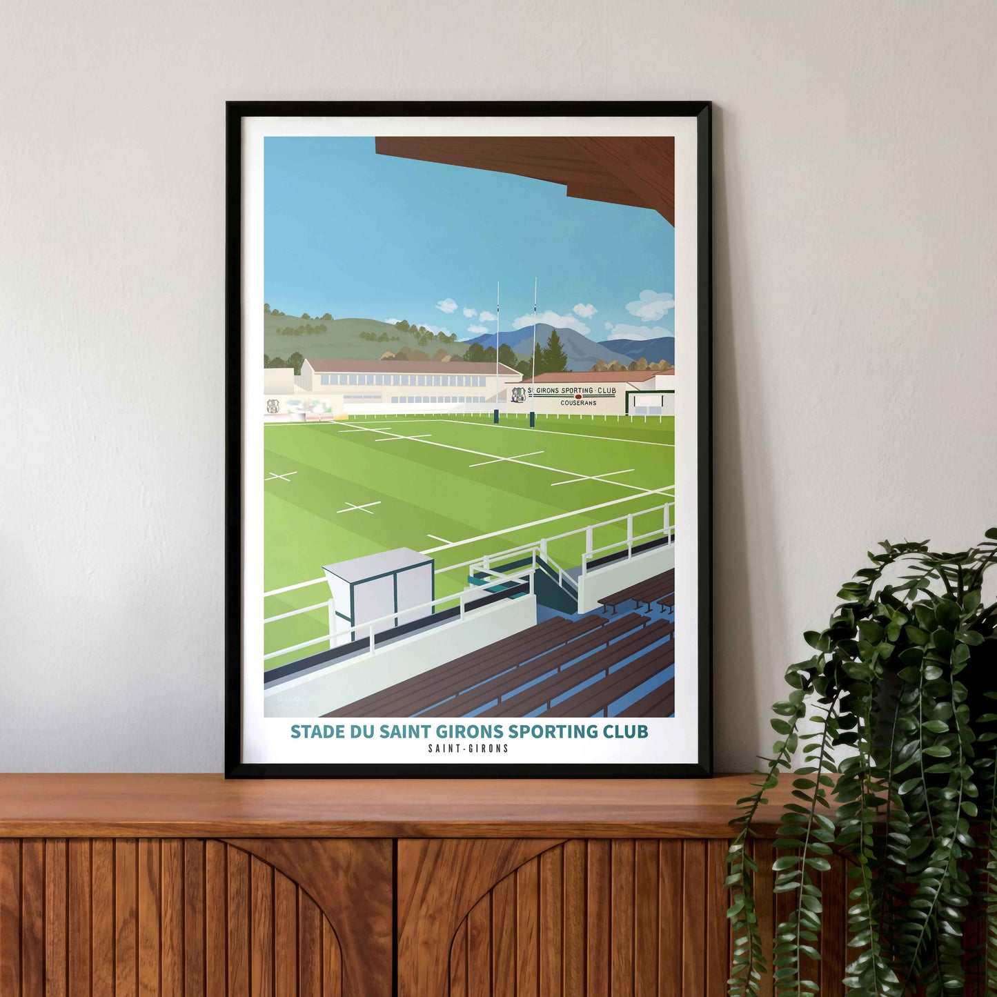 Illustration du stade de rugby de Saint-Girons dans un cadre noir, posée sur un buffet vintage en bois avec une plante verte.