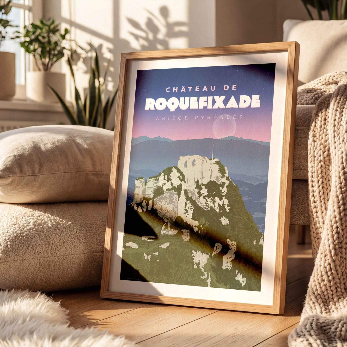 Affiche décorative de Roquefixade dans un cadre en bois, posée sur un tapis blanc épais près d'un coussin moelleux.