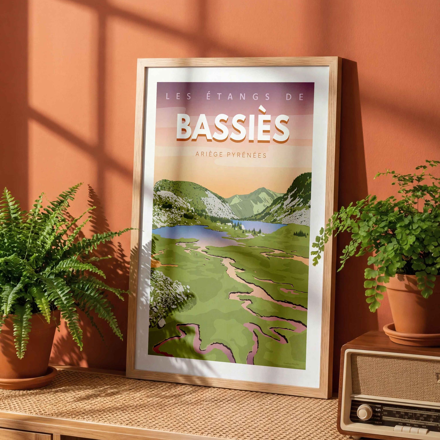 Affiche Bassiès encadrée, posée sur une étagère en bois devant un mur de couleur terracotta avec des plantes vertes.