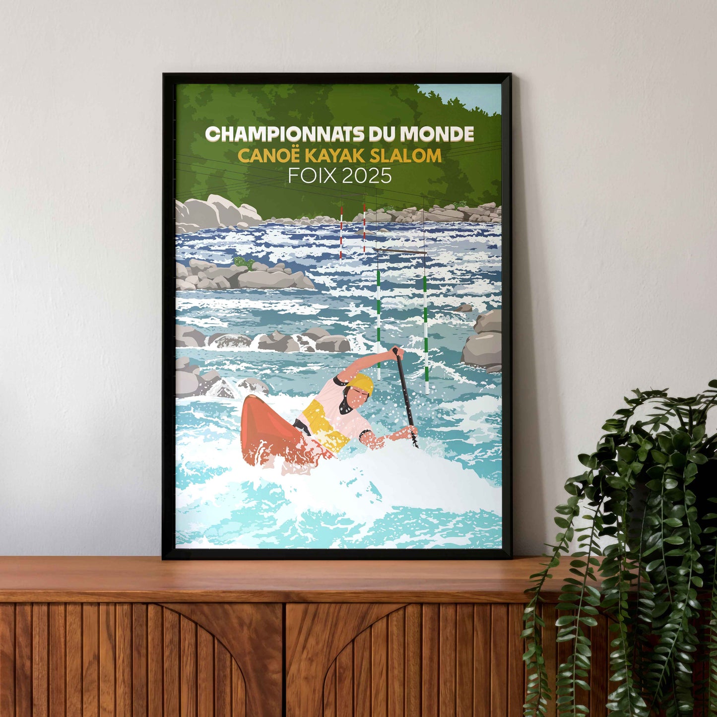 Affiche commémorative Foix 2025 de canoë-kayak dans un cadre noir élégant, posée sur un buffet en bois sombre avec une plante verte décorative.