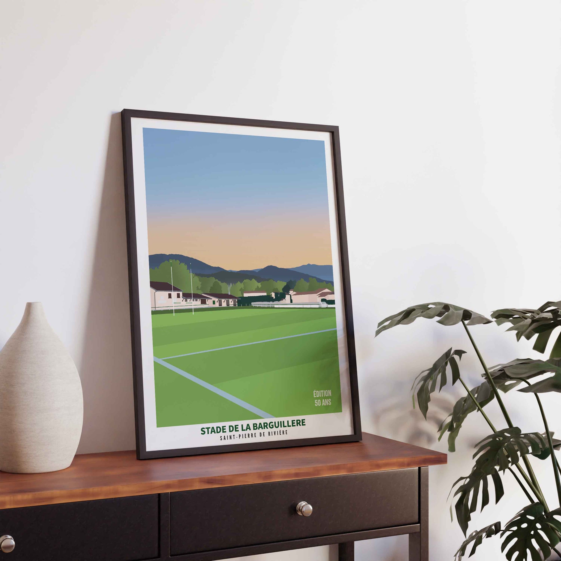 Illustration du stade de rugby de la Barguillère dans un cadre noir fin, posée sur une console en bois sombre à côté d'un vase.