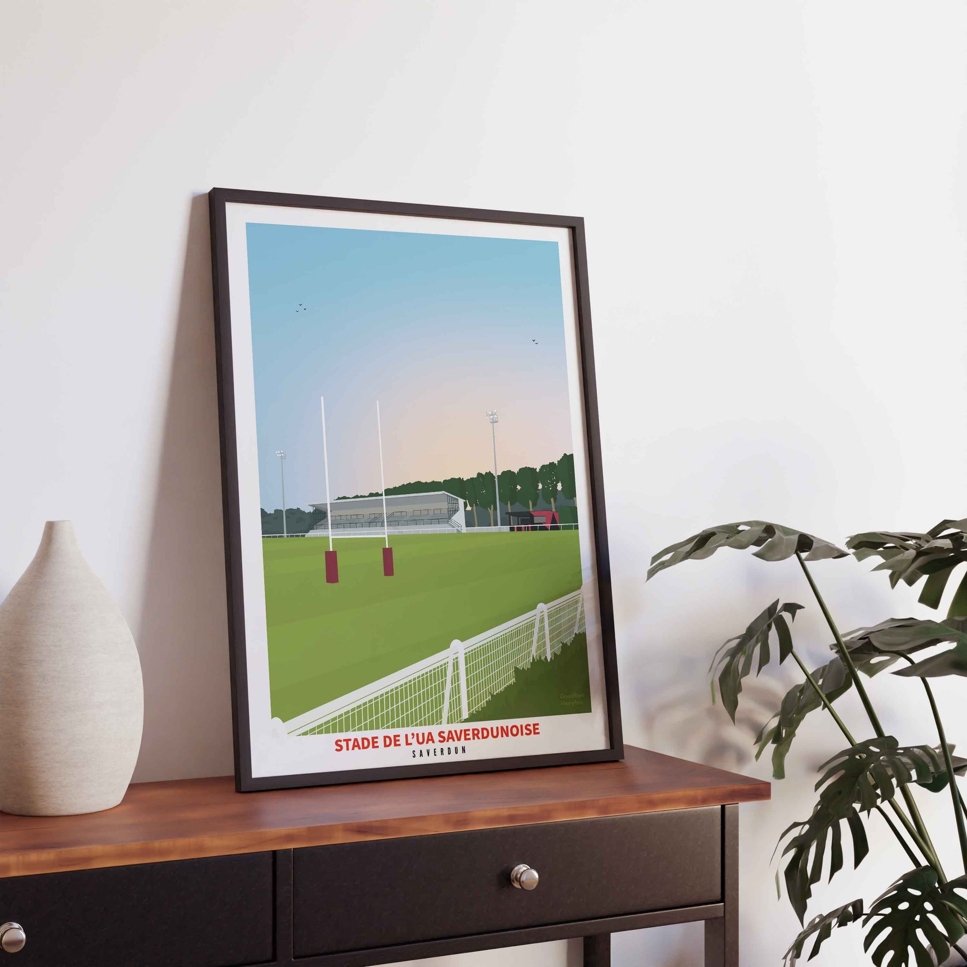 Illustration du stade Paul Fines de Saverdun dans un cadre noir fin, posée sur une console en bois sombre à côté d'un vase blanc.
