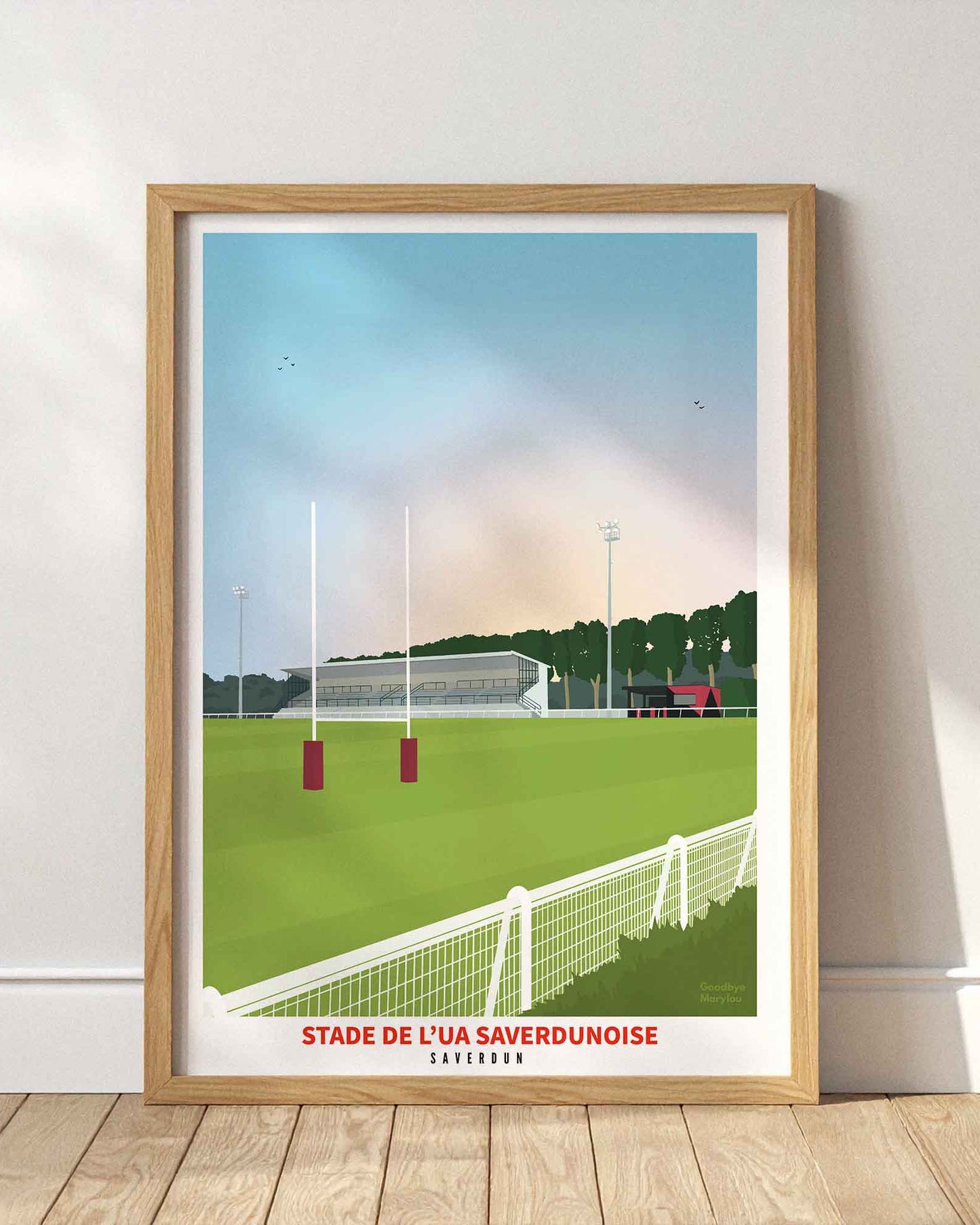 Affiche du stade de l’UA Saverdunoise (Saverdun) encadrée en bois clair, posée au sol contre un mur blanc épuré avec des jeux d'ombres.